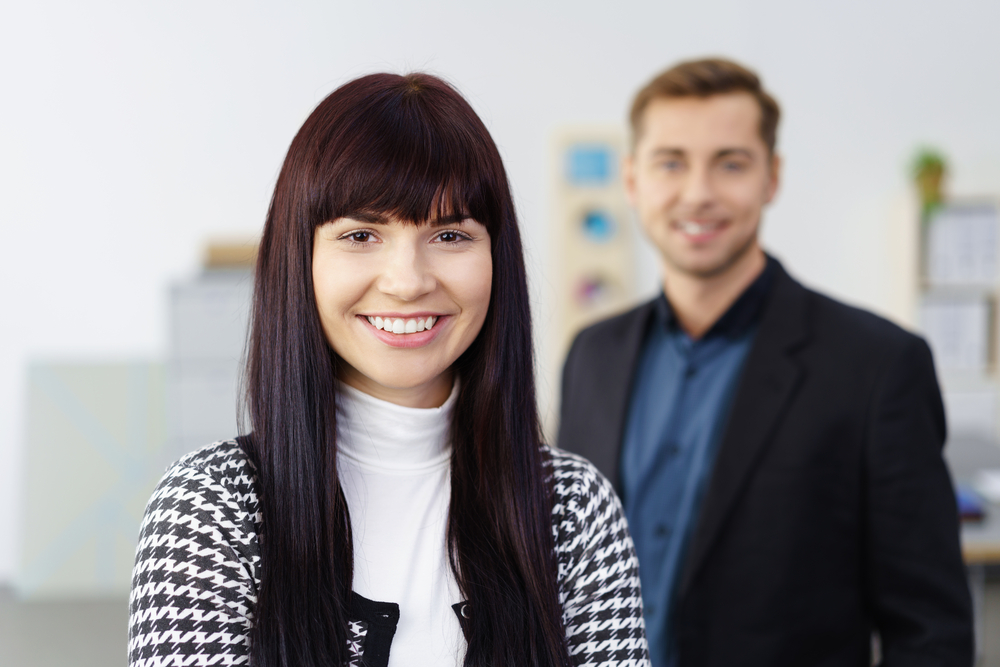 Happy confident business manageress or entrepreneur posing looking at the camera with a friendly smile with a male co-worker behind